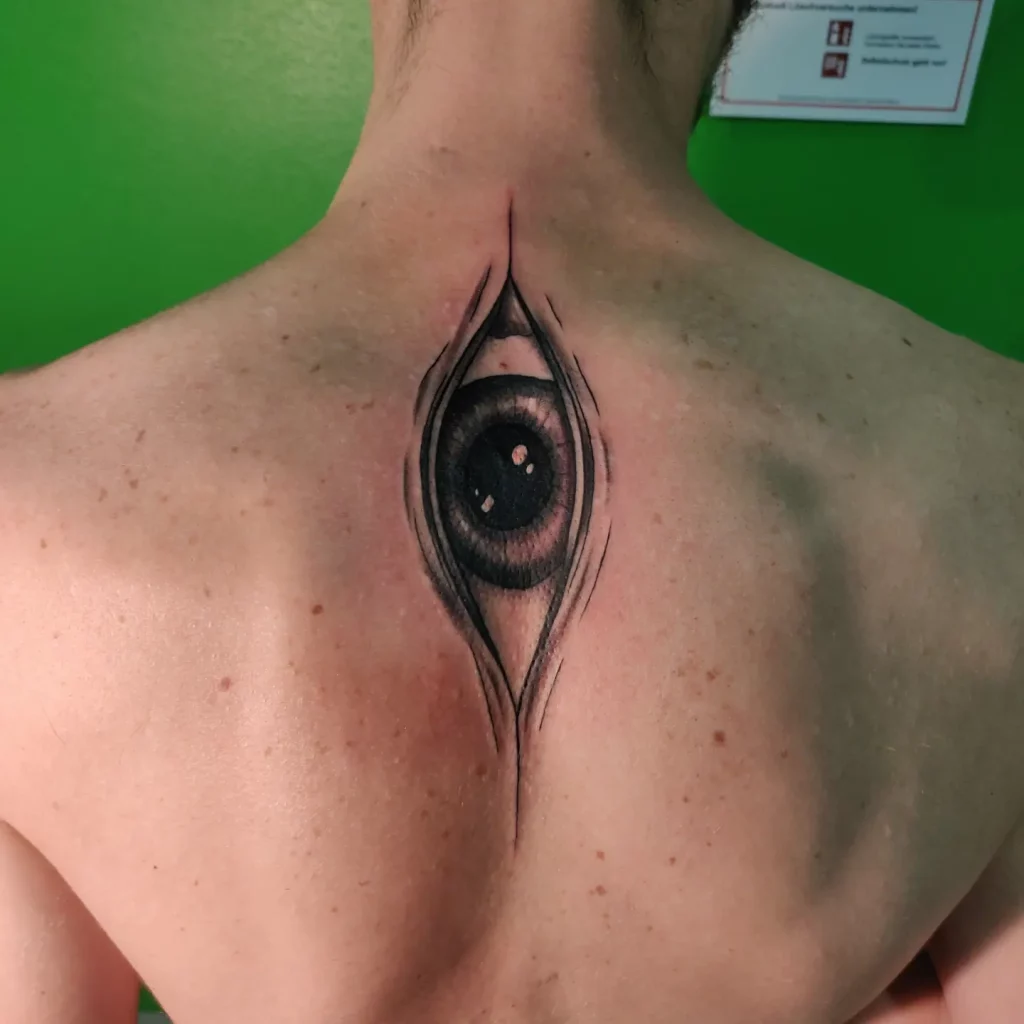 a tattoo on the upper back. the design is realism in black and grey and the tattoo is showing a vertical eye coming out of the back.