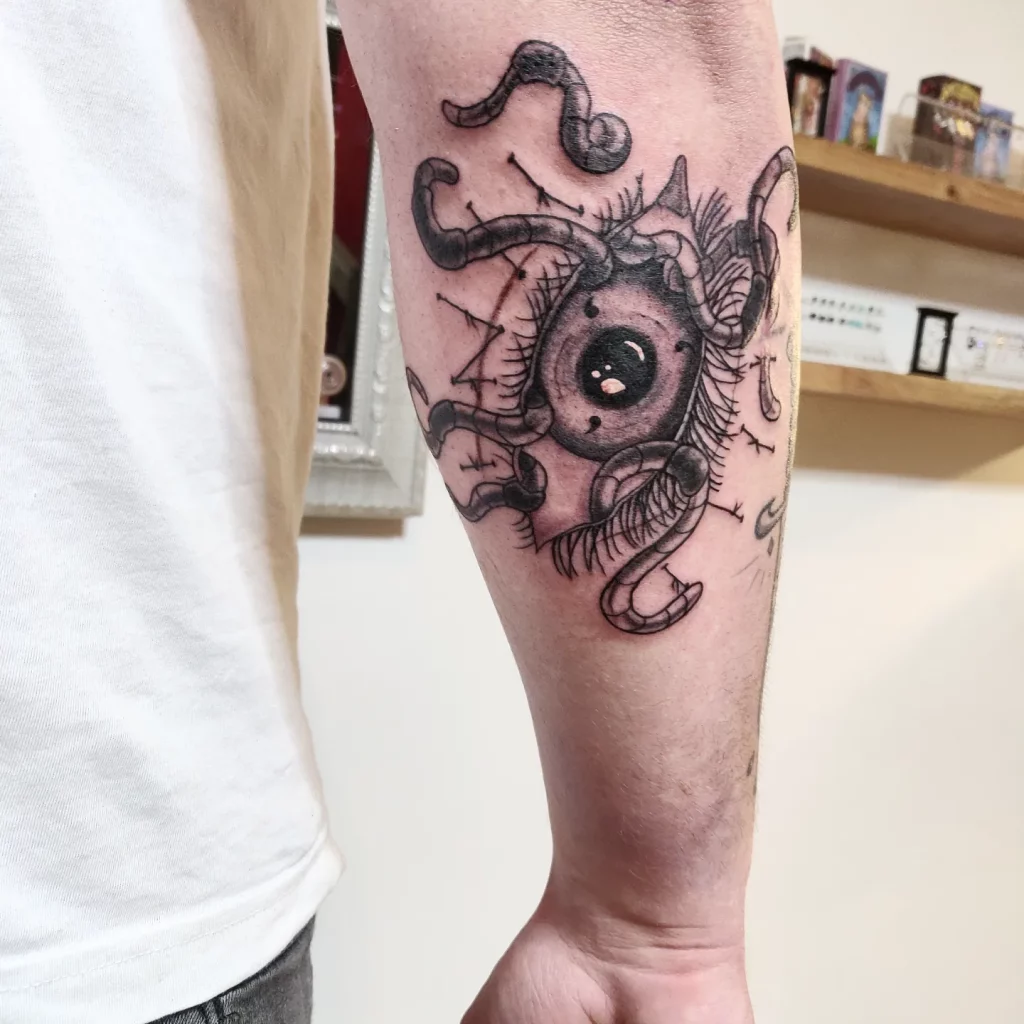 a black and grey tattoo on the back of the lower arm. It shows a big sharingan eye, worms are crawling out.
