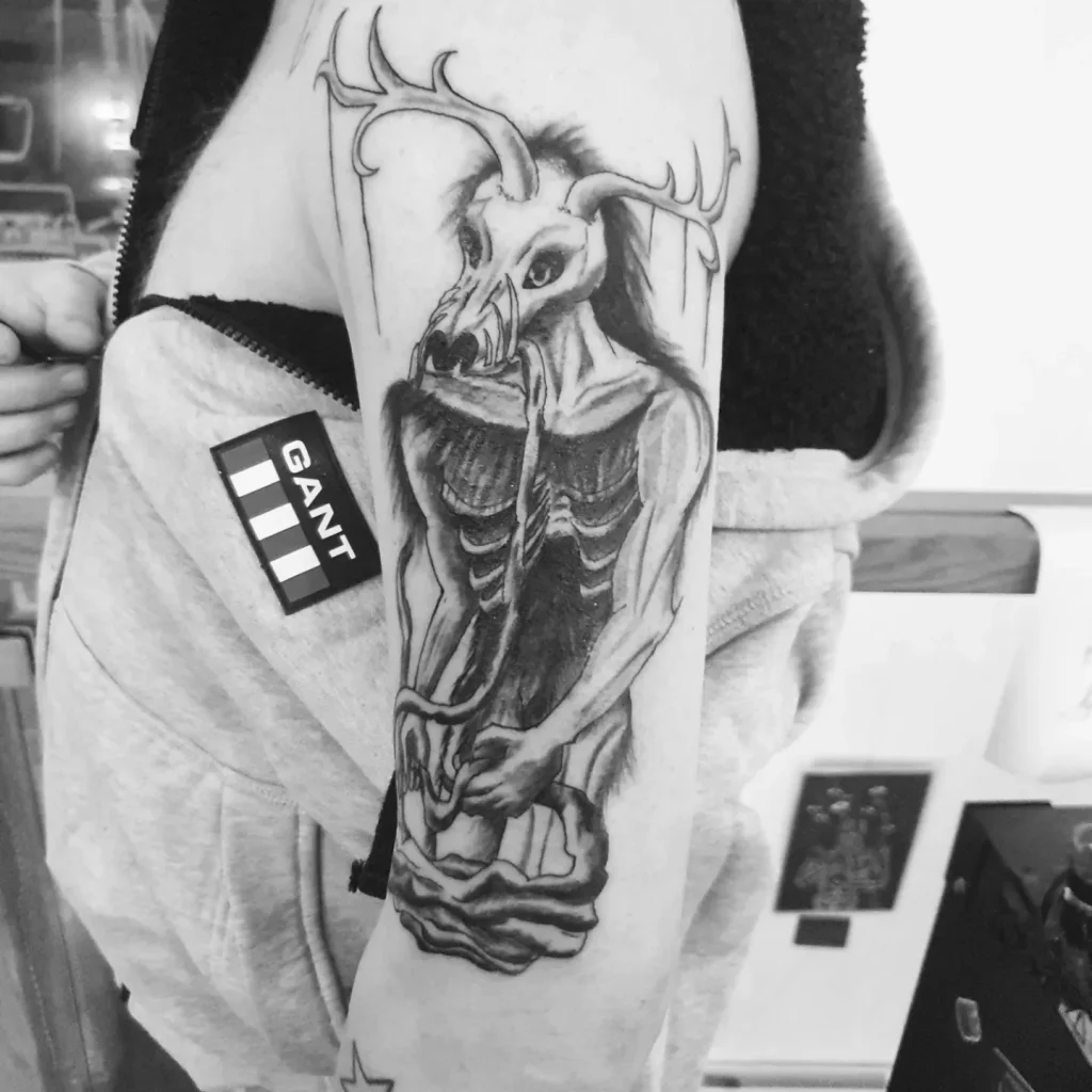 upperarm tattoo of a wendigo, eating some human flesh and a long piece of a gut in its claws and mouth, black and grey