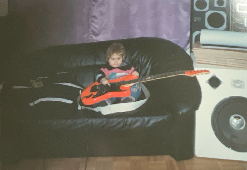 3 year old lucy sitting on a black sofa playing a red electric guitar