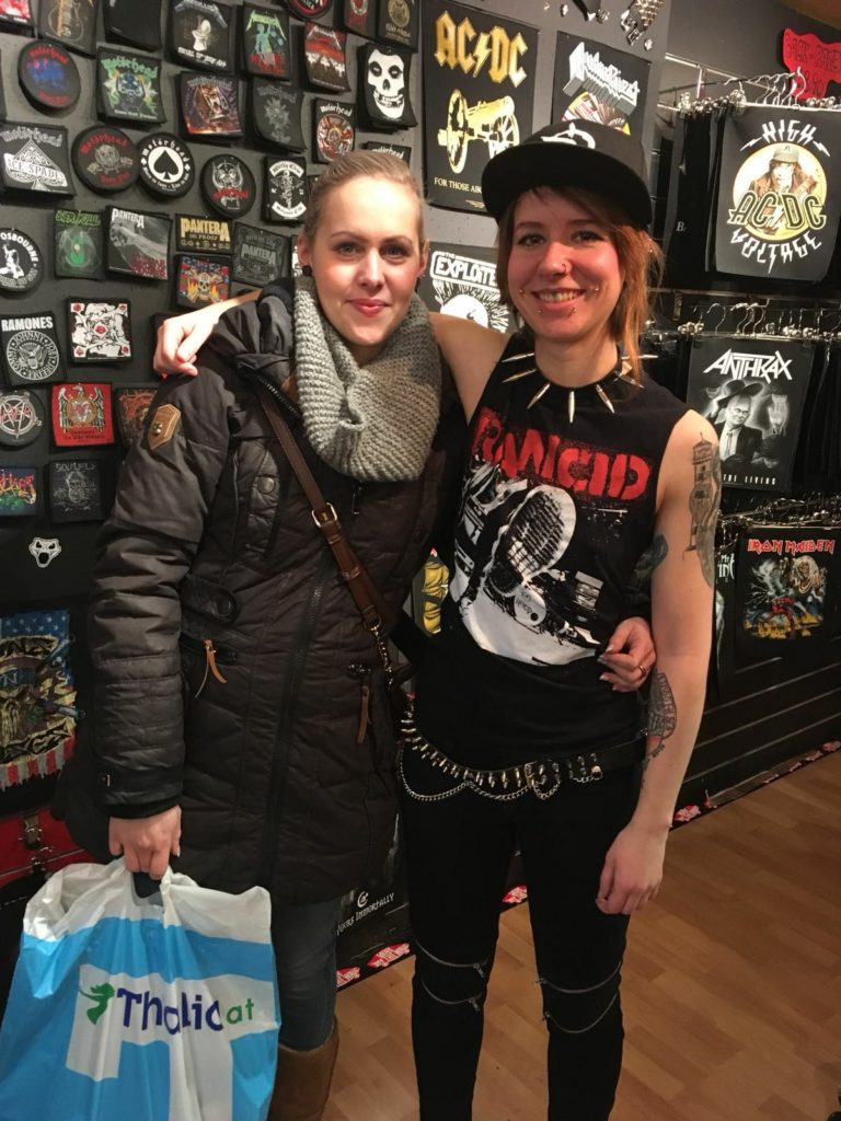 Lucy with her client arm in arm facing the camera at the shop "Rattlesnake" in front of the wall decorated with patches. Lucy is wearing a "Rancid" shirt and a spiked necklace